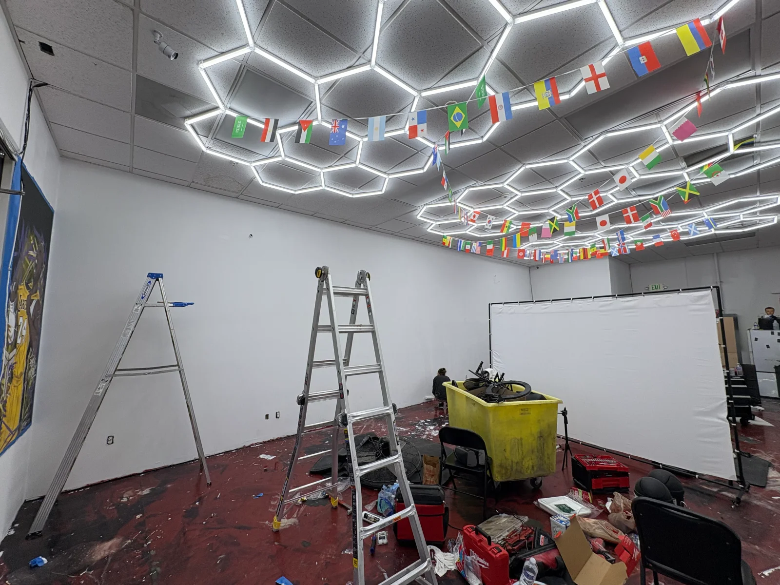 Open commercial space with hexagonal LED ceiling lights, white painted walls, white backdrop, and aluminum ladders during buildout