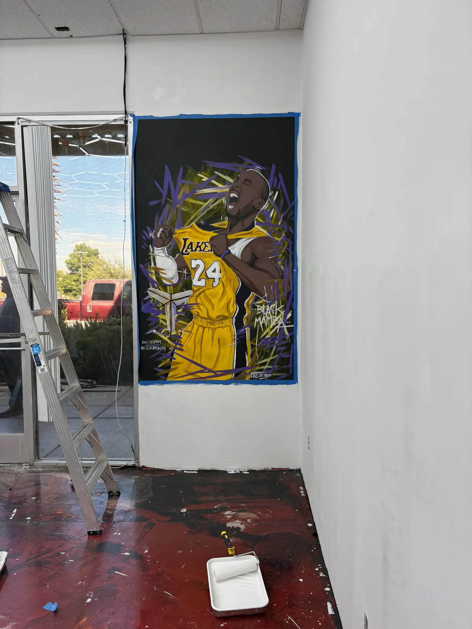 Hand-painted basketball player mural in gold jersey with purple brushwork on white interior wall, taped edges and paint tray on floor