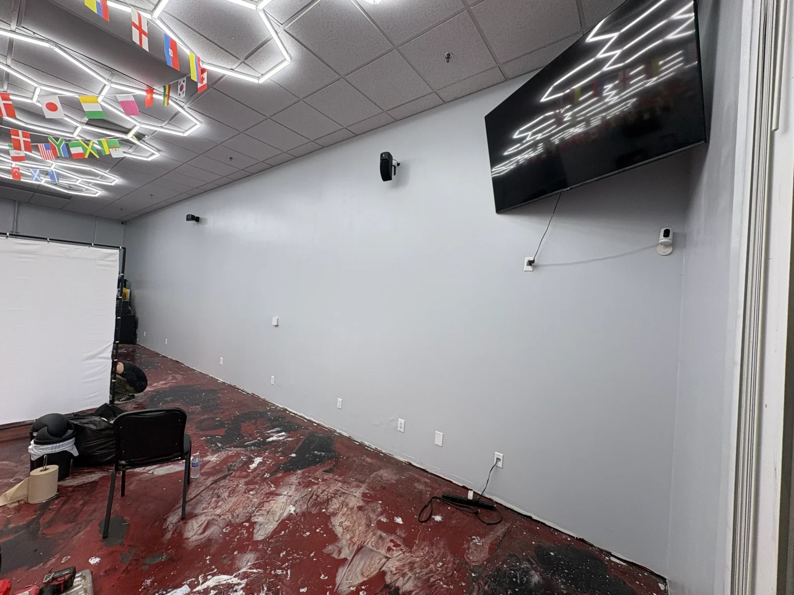 Freshly painted light gray interior wall with mounted TV, wall outlets, and paint-splattered concrete floor during finishing work