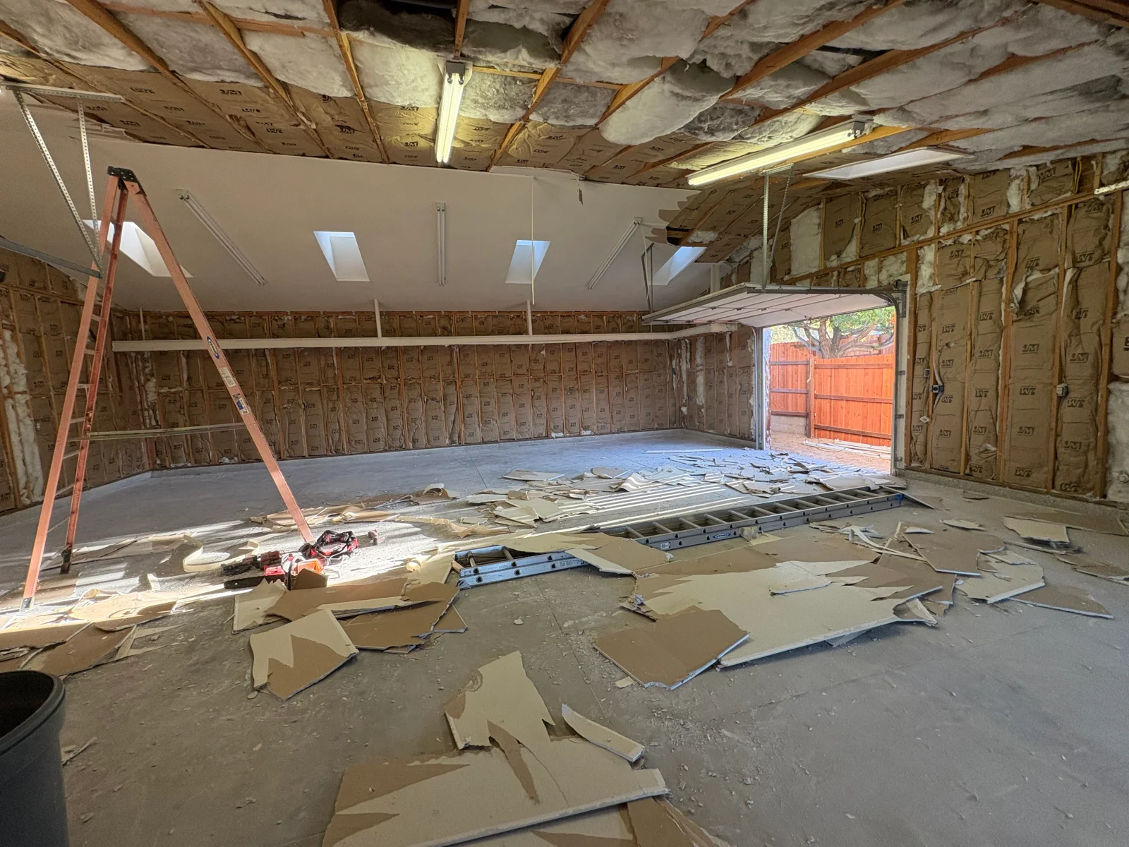 Garage interior with drywall removed, exposed wood studs, pink and yellow batt insulation, and gypsum debris on concrete floor