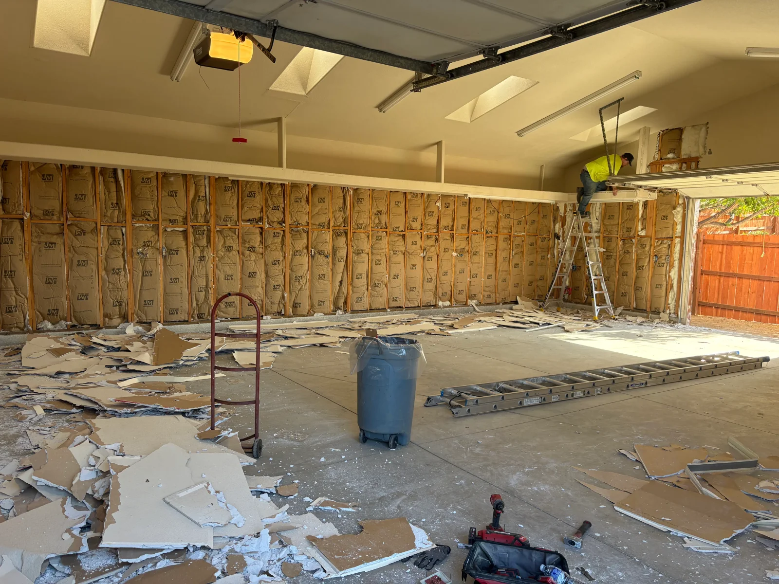 Garage interior with drywall removed, exposed wood studs and fiberglass insulation batts, debris on concrete floor, extension ladder and cordless drill