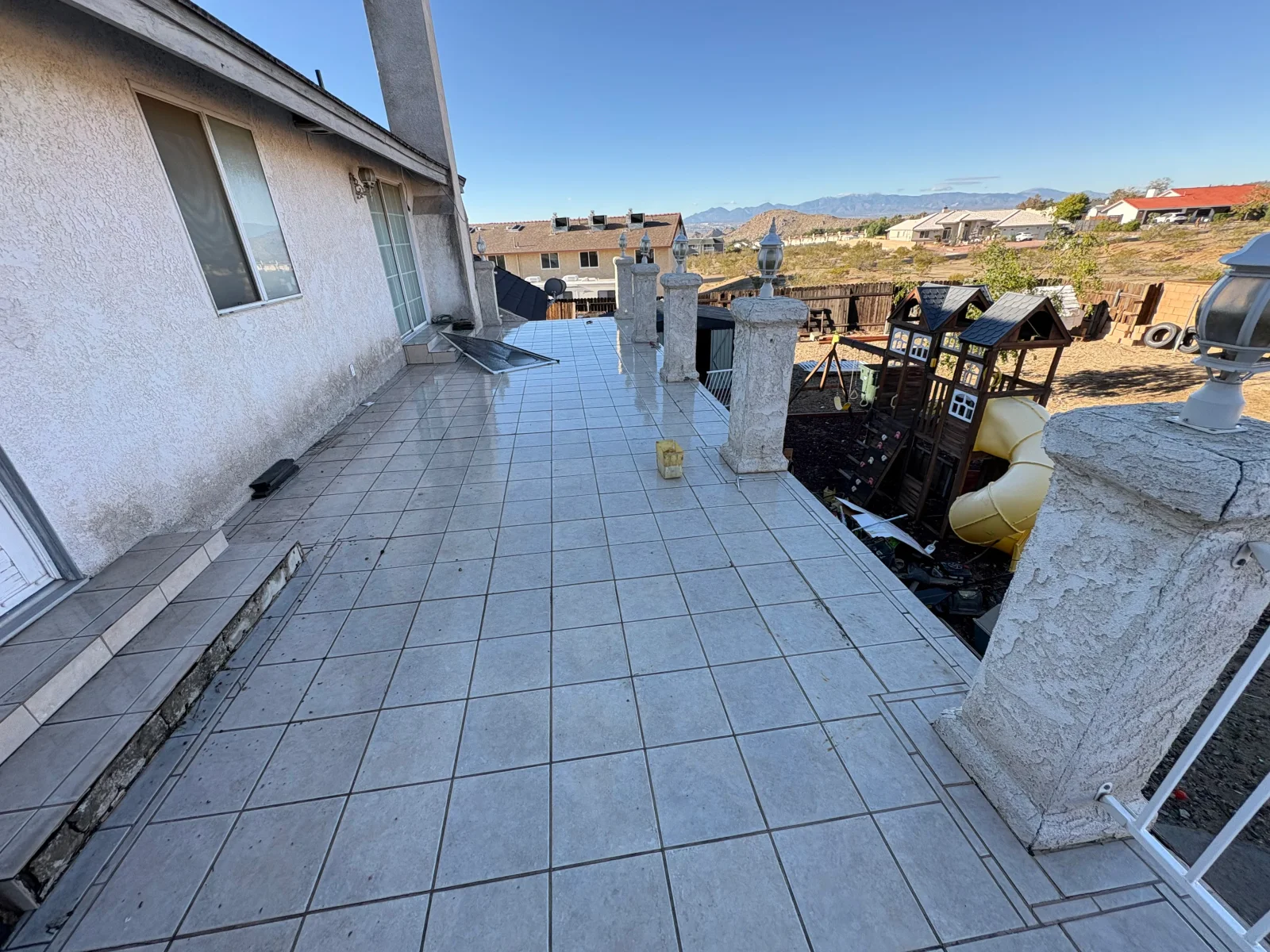 Glazed gray square tile patio with dark grout, stucco pillars with lantern lights, and stucco exterior wall