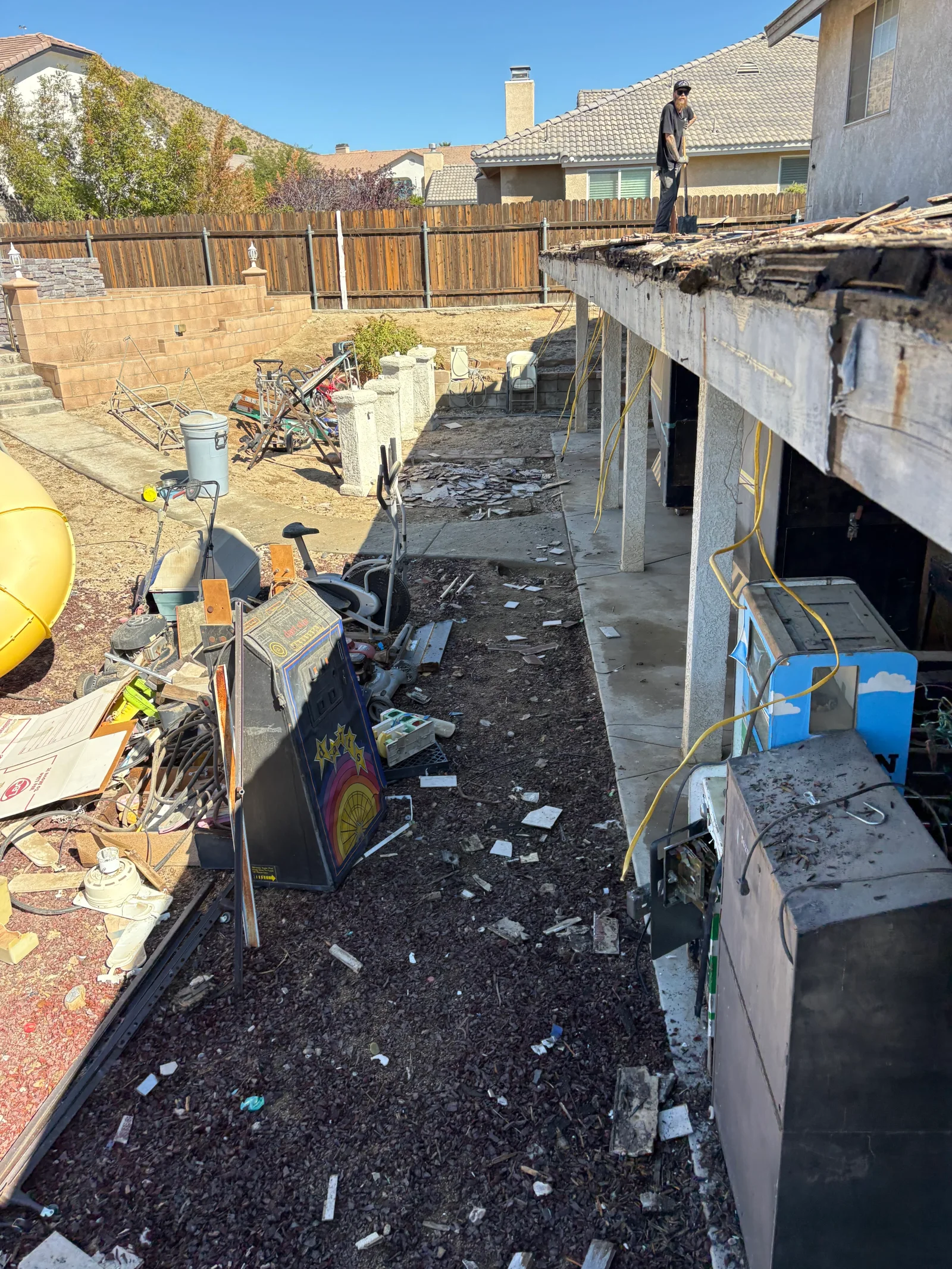 Backyard with damaged patio roof partially stripped, arcade cabinets, exercise equipment, and debris piled for removal