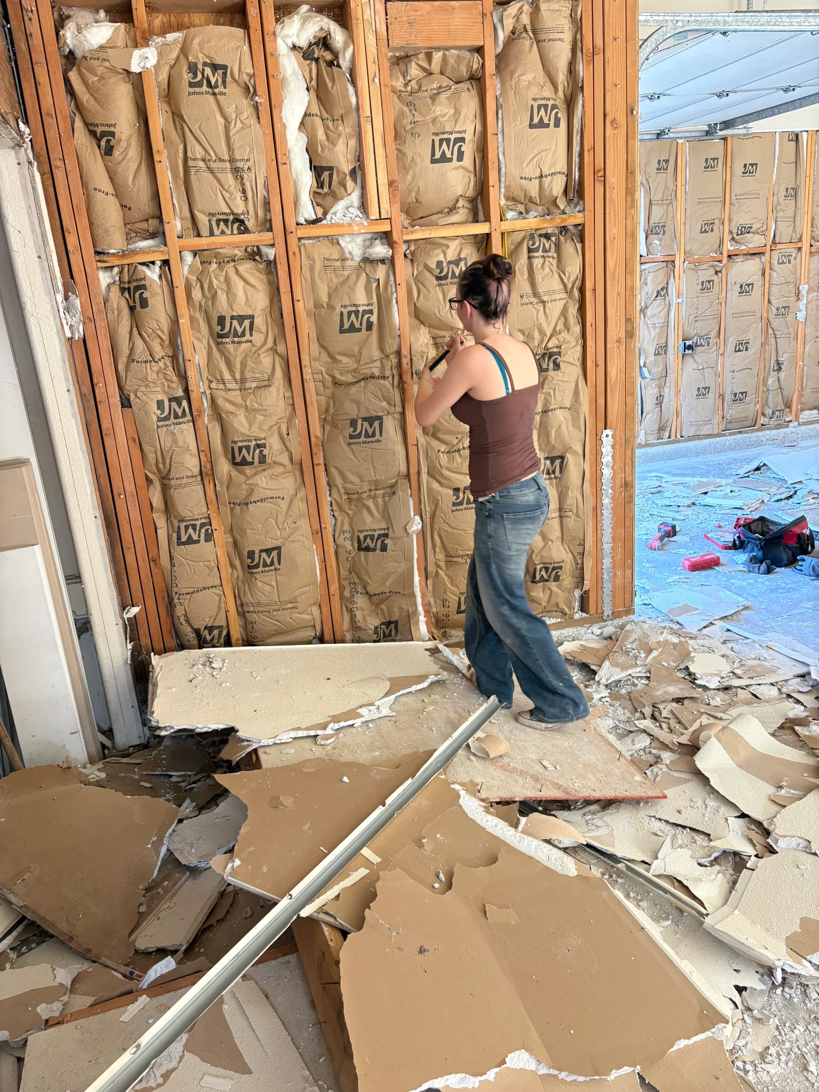 Worker marking wood-stud wall filled with Johns Manville kraft-faced fiberglass insulation batts during drywall removal