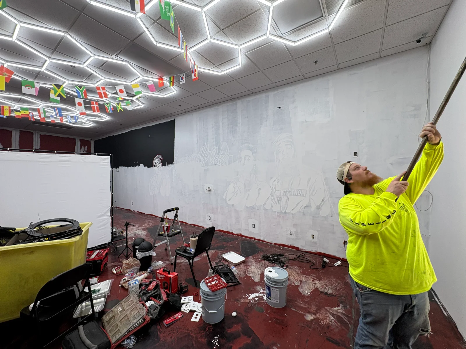 Worker rolling primer on a large interior wall with a sketched mural layout, paint buckets and tools on a splattered floor.
