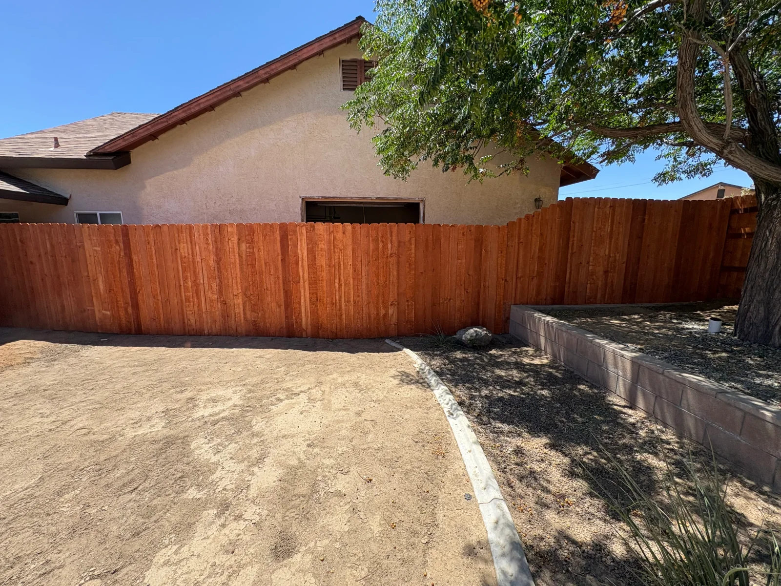 Stained wood privacy fence with concrete edging beside stucco home, gravel planter with block wall