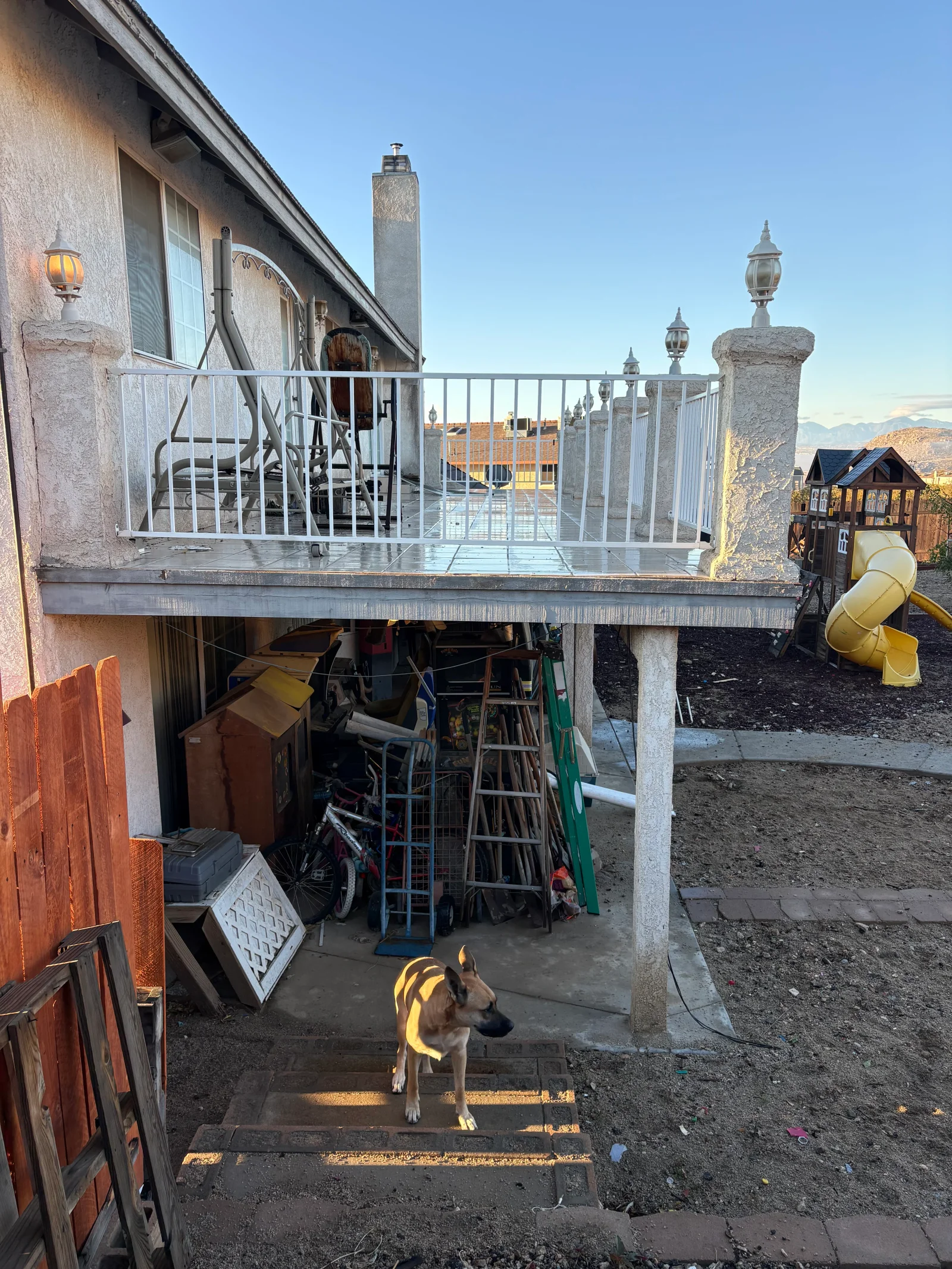 Raised patio deck with white metal railing, stucco pillars, and tile surface above a covered yard area