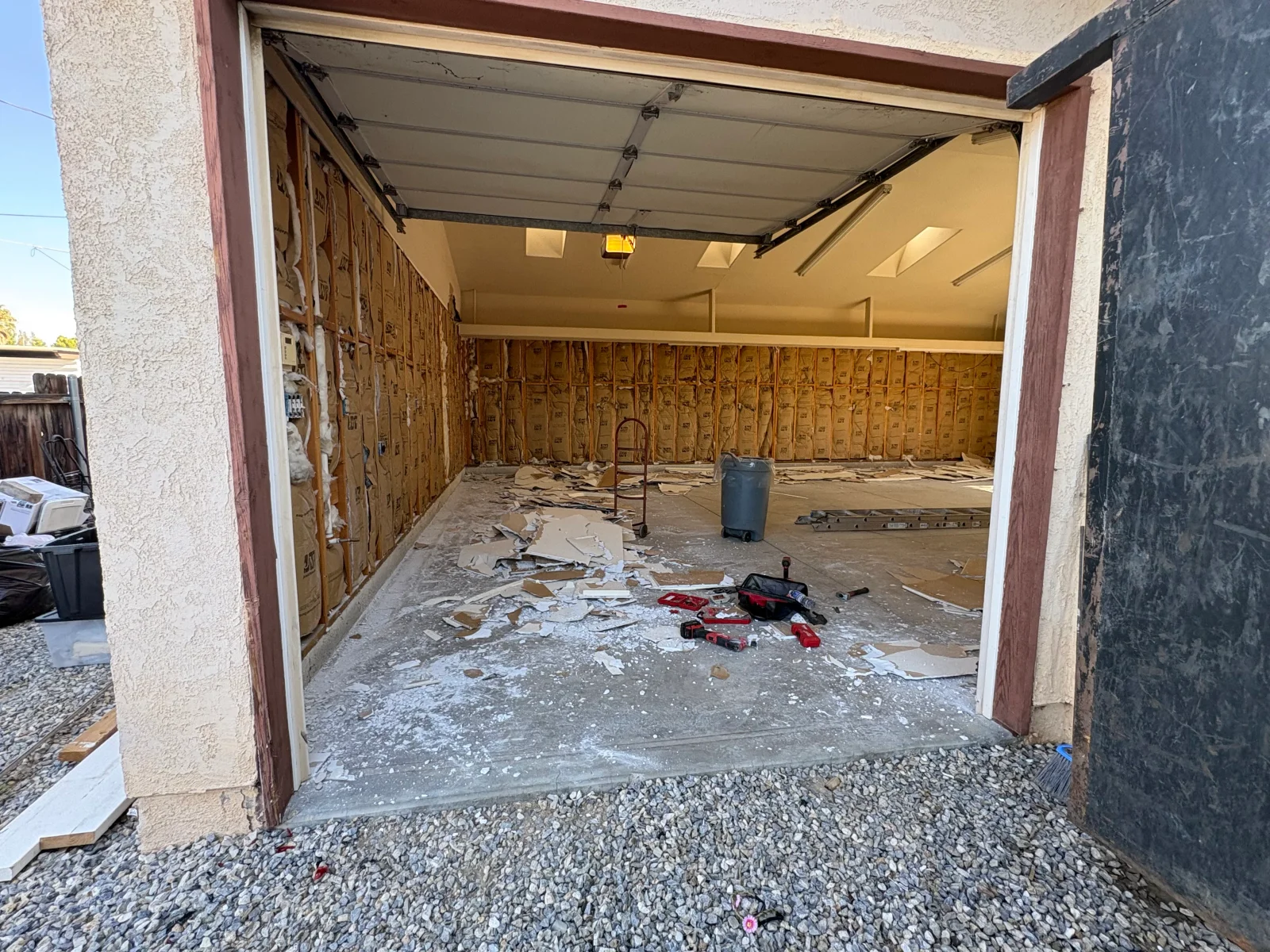 Garage interior with drywall removed, exposed insulation batts and wood framing, debris on concrete floor, and tools scattered near an open garage door