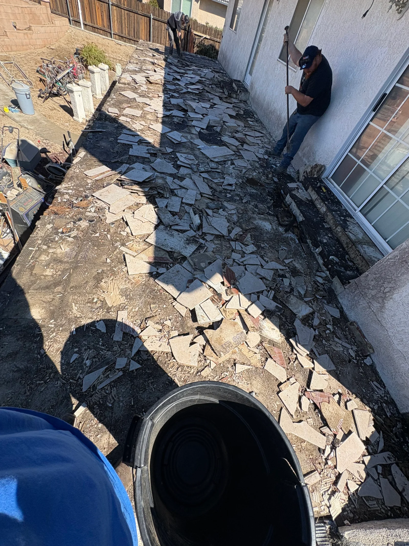 Crew demolishing tile flooring beside stucco wall, broken tile and mortar debris over concrete slab, black debris bucket in foreground