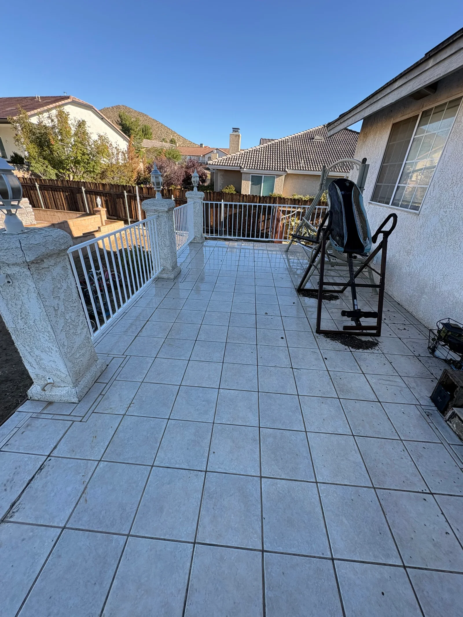 Large light gray square tile patio with dark grout lines, stucco pillar, white metal railing, and stucco house wall