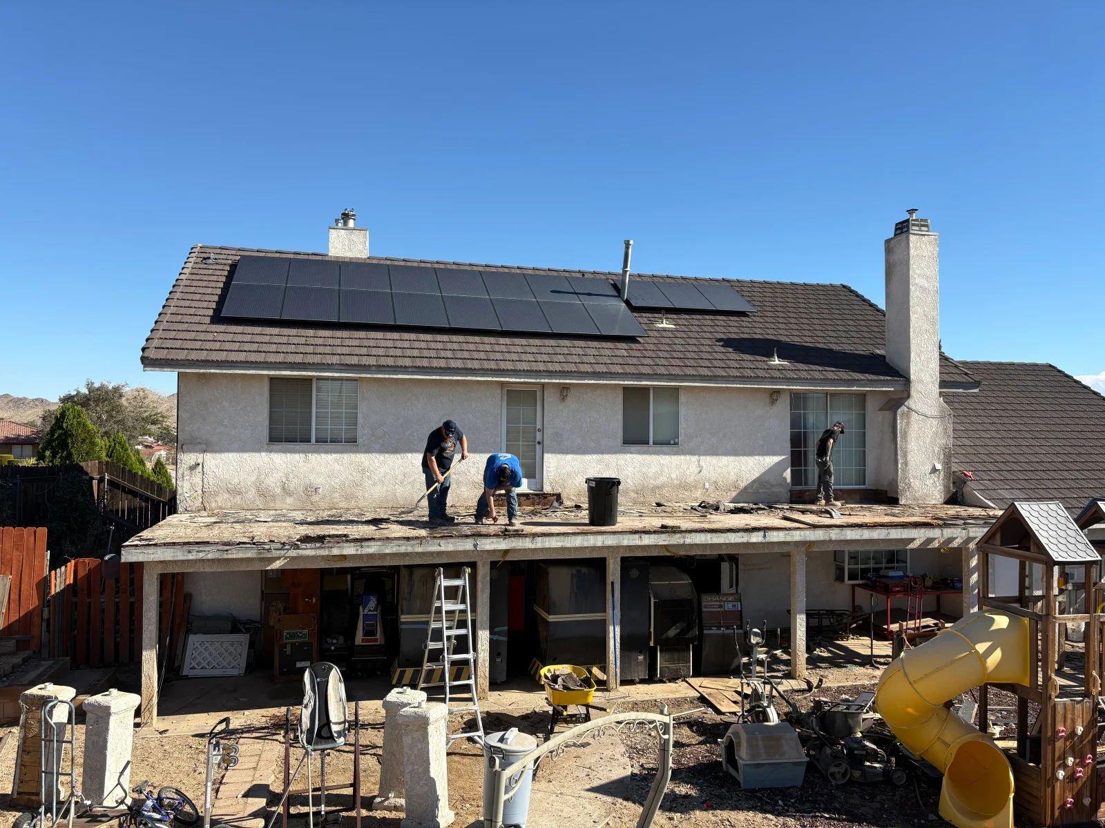Workers demolishing a second-story deck surface with pry bars, exposed stucco-finished substructure, and debris bin on site