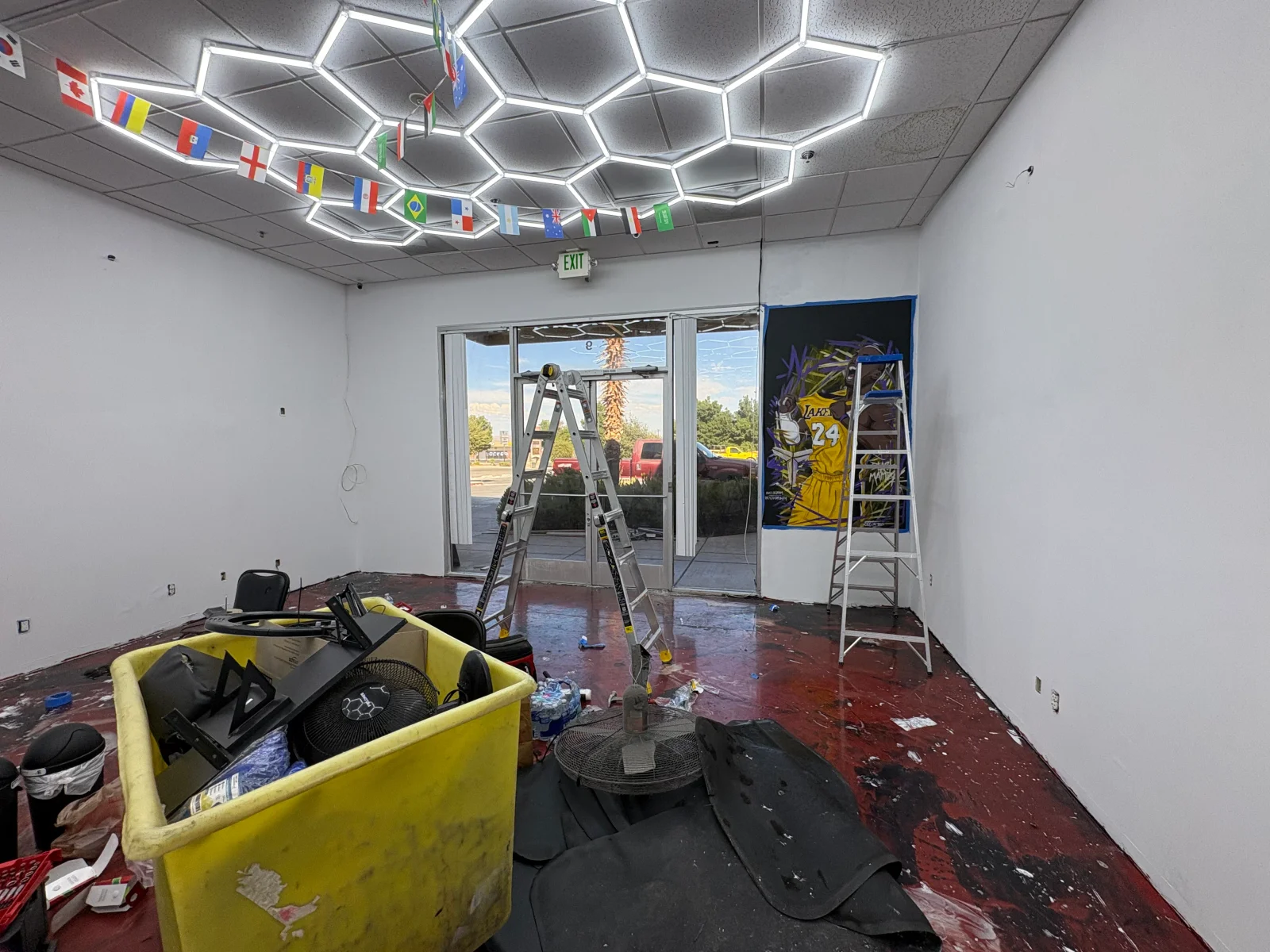 Empty commercial interior with hexagonal LED ceiling lights, white painted walls, red floor, glass entry doors, and a mural on the wall