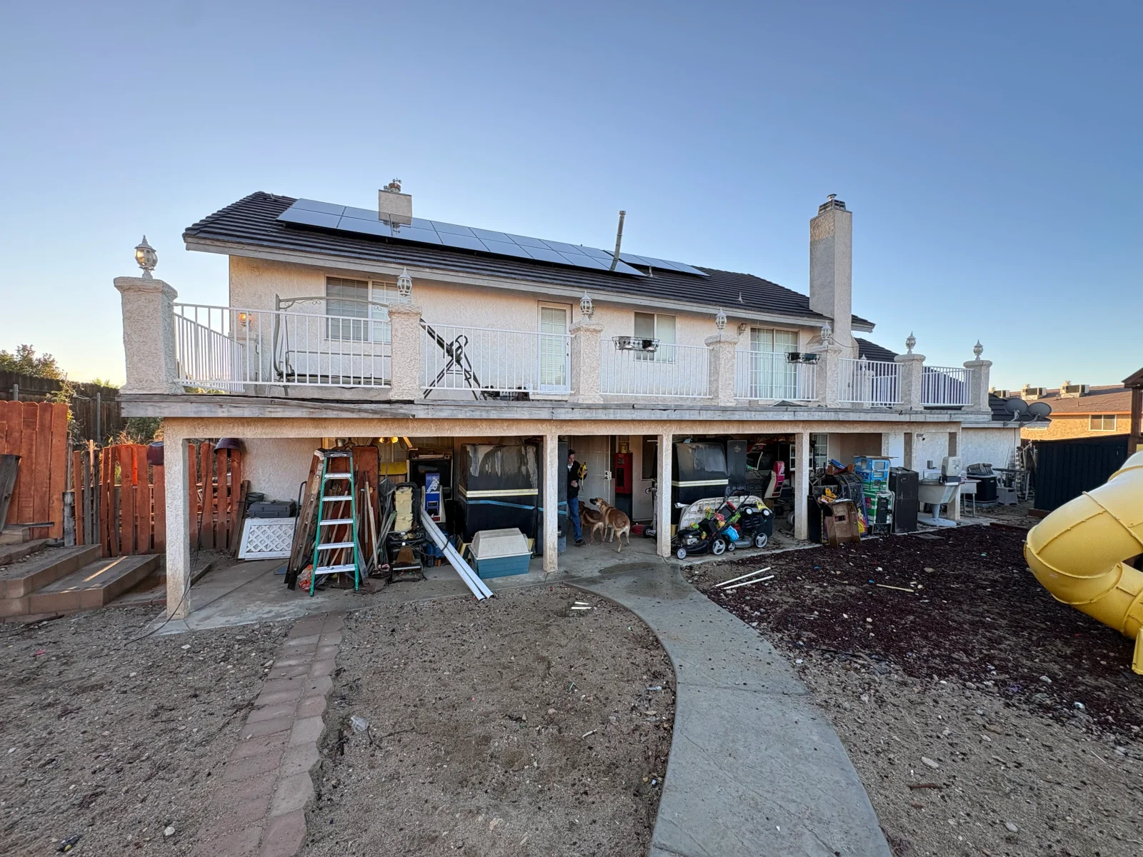 Stucco two-story home with solar panels and white balcony railings, backyard cluttered with furniture, appliances, and lumber