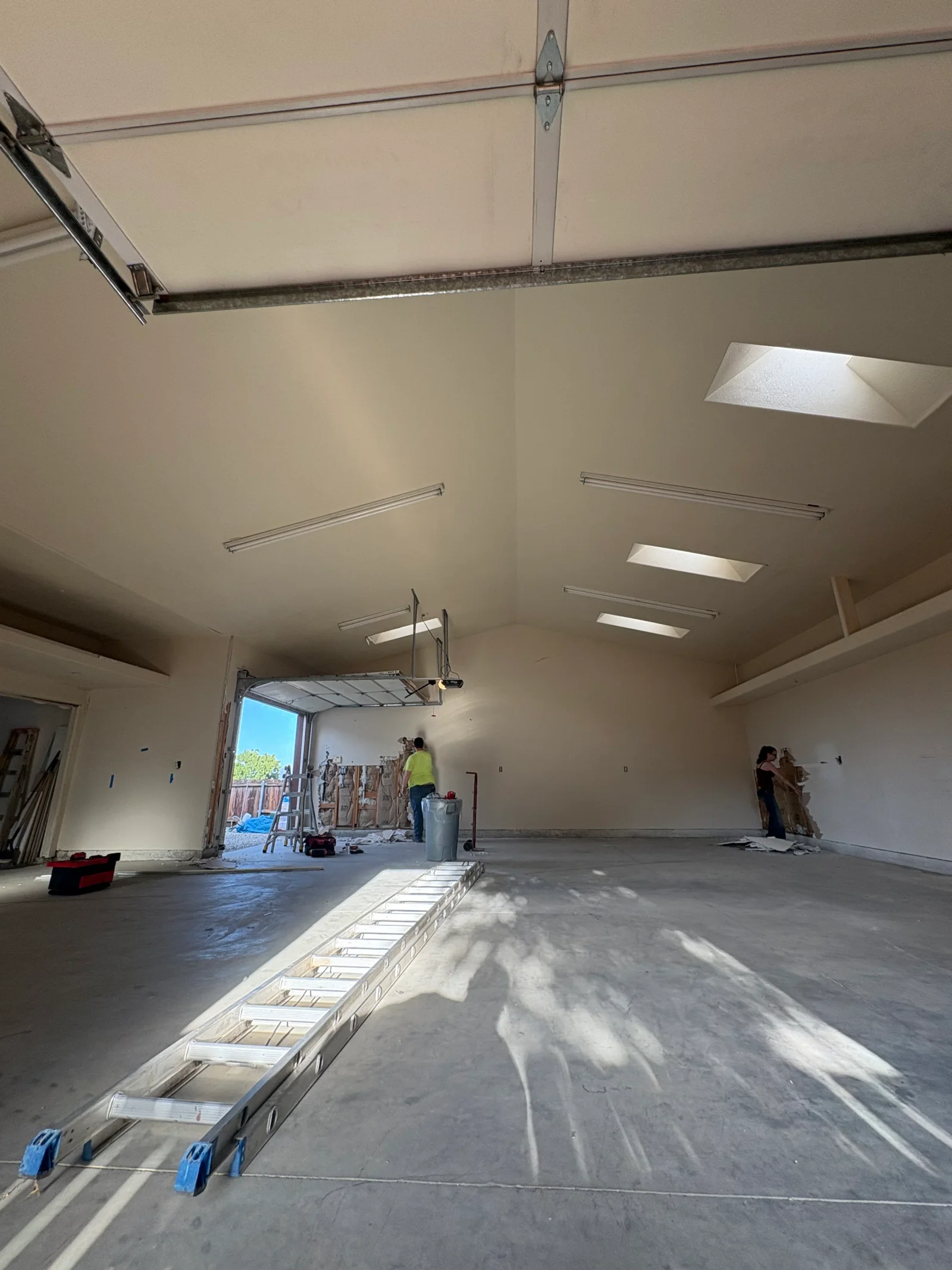 Garage interior with drywall removed from walls exposing wood framing and insulation, aluminum extension ladder on concrete floor, skylights overhead