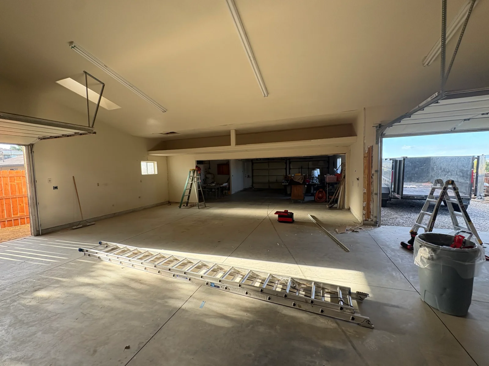 Gutted two-car garage with bare drywall, exposed framing, concrete floor, aluminum ladders, and open overhead doors
