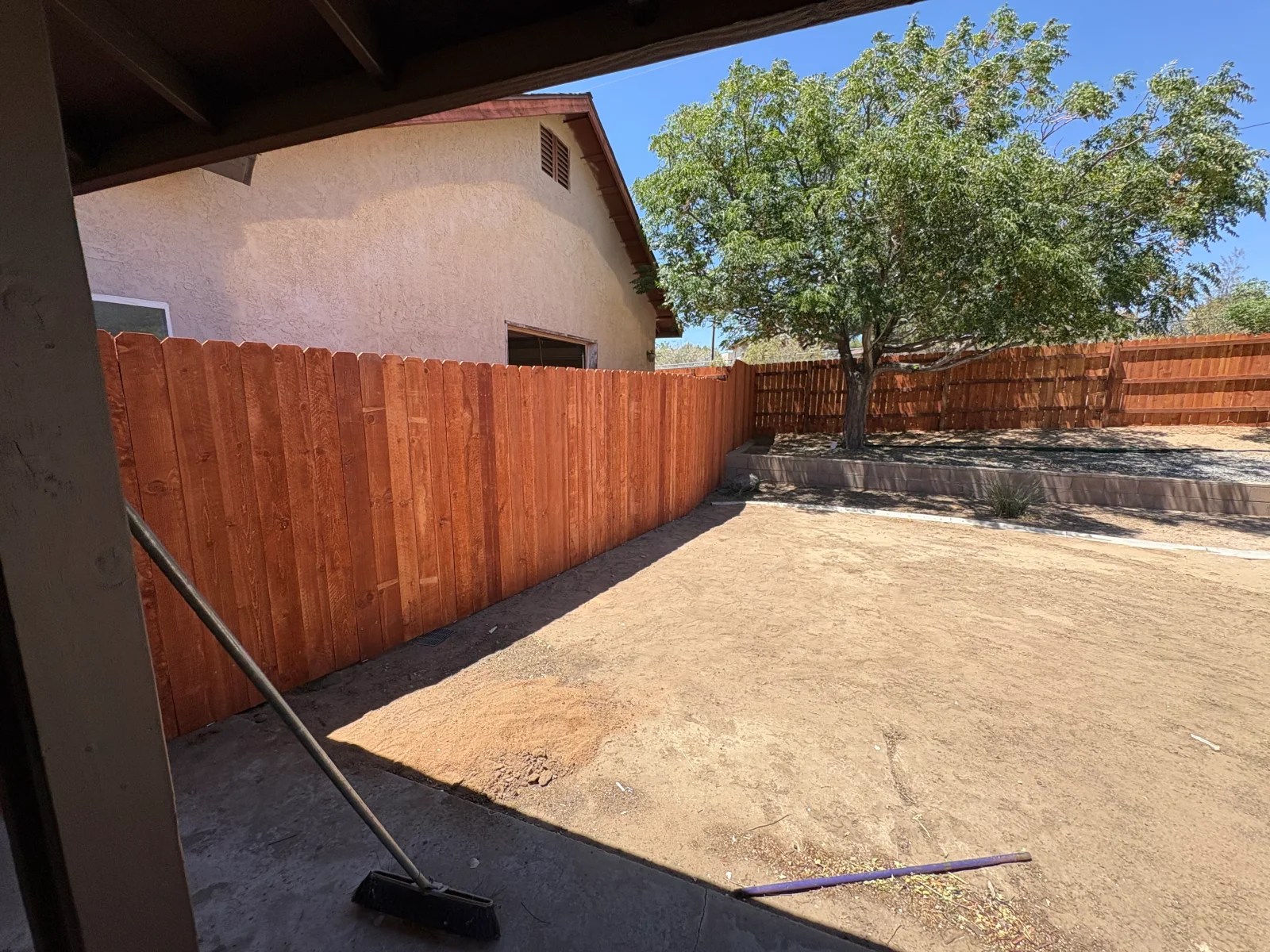 Stained wood privacy fence with scalloped top beside stucco home, bare dirt yard, and shade tree