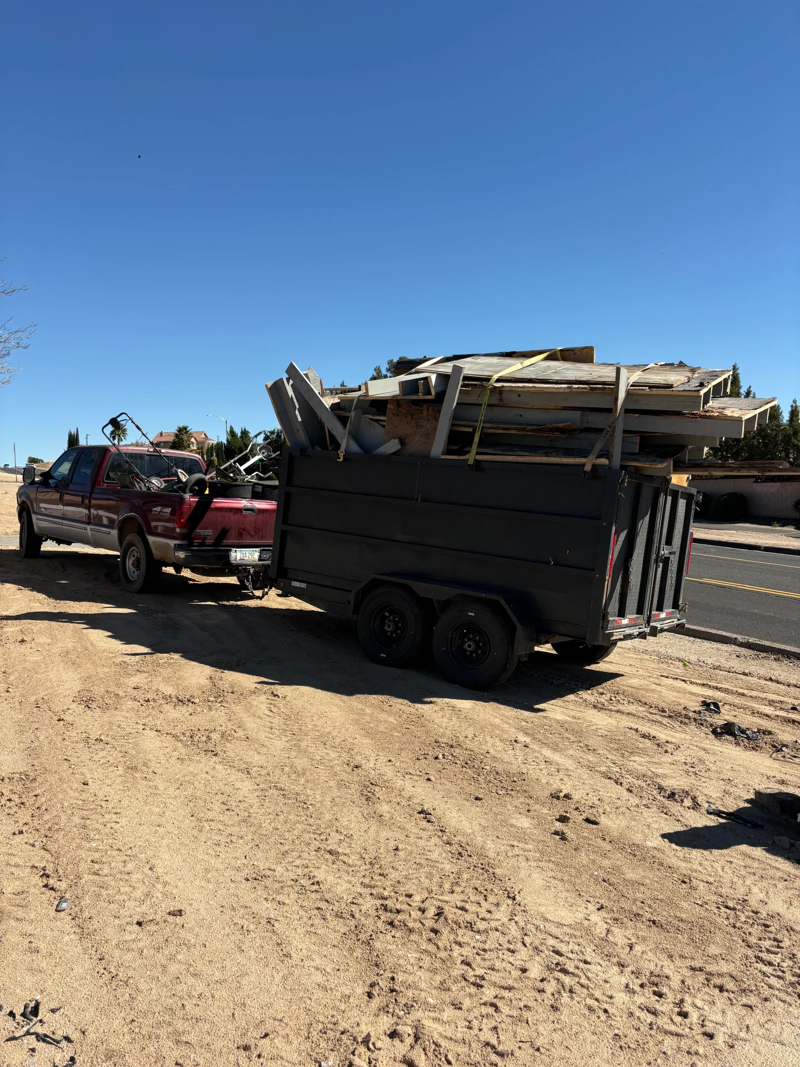 Black dump trailer loaded with plywood, lumber, and framing debris secured by yellow straps, towed by a red pickup truck