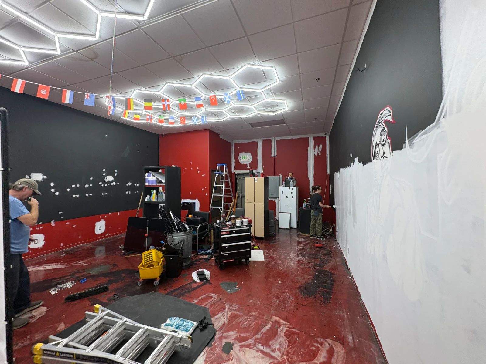 Interior commercial space with white primed wall, sketched mural outline, charcoal and red painted walls, and hexagonal LED ceiling lights