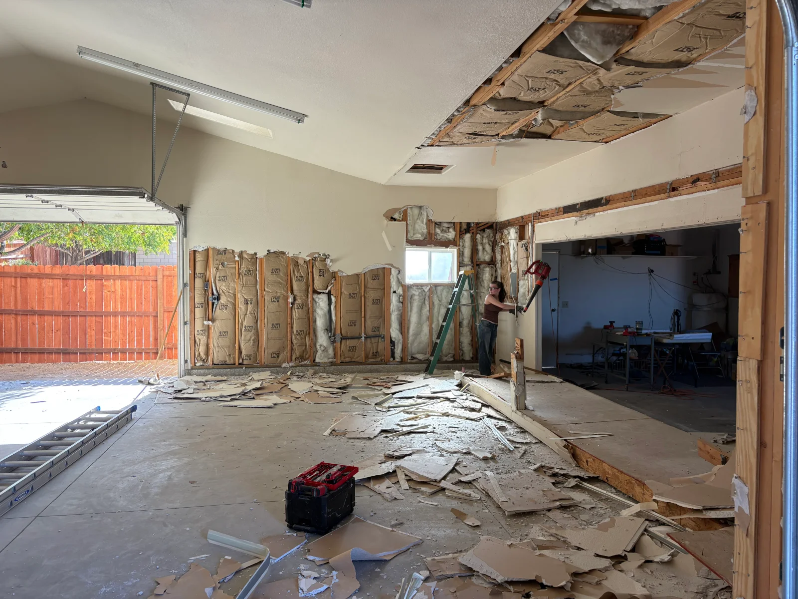 Garage interior demolition showing exposed wall studs, batt insulation, removed drywall debris on concrete floor, and a worker on a step ladder